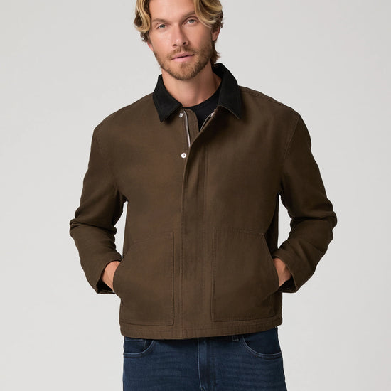 A man wearing a dark brown waxed canvas utility jacket with a black collar, paired with blue jeans, photographed against a solid light gray background.