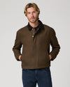 A man wearing a dark brown waxed canvas utility jacket with a black collar, paired with blue jeans, photographed against a solid light gray background.