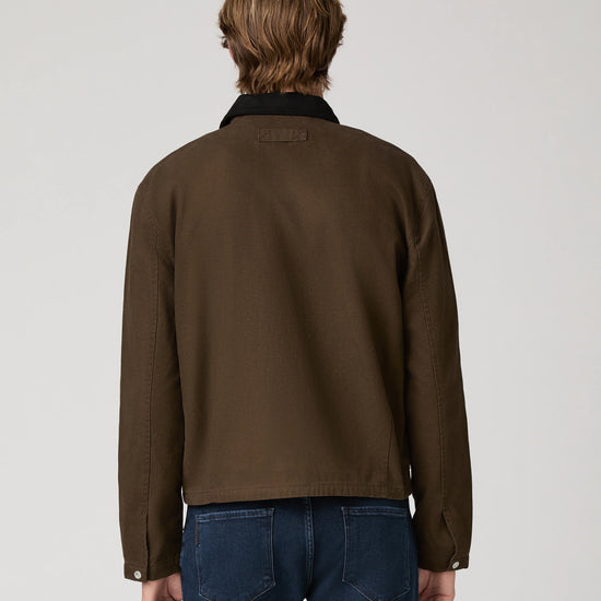 A man viewed from behind wearing a dark brown utility jacket with a black collar and blue jeans, set against a plain light gray studio background.