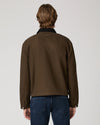 A man viewed from behind wearing a dark brown utility jacket with a black collar and blue jeans, set against a plain light gray studio background.