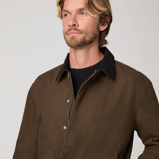 A man wearing a brown corduroy jacket with a black corduroy collar over a black t-shirt, photographed against a neutral light gray background.