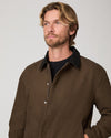 A man wearing a brown corduroy jacket with a black corduroy collar over a black t-shirt, photographed against a neutral light gray background.