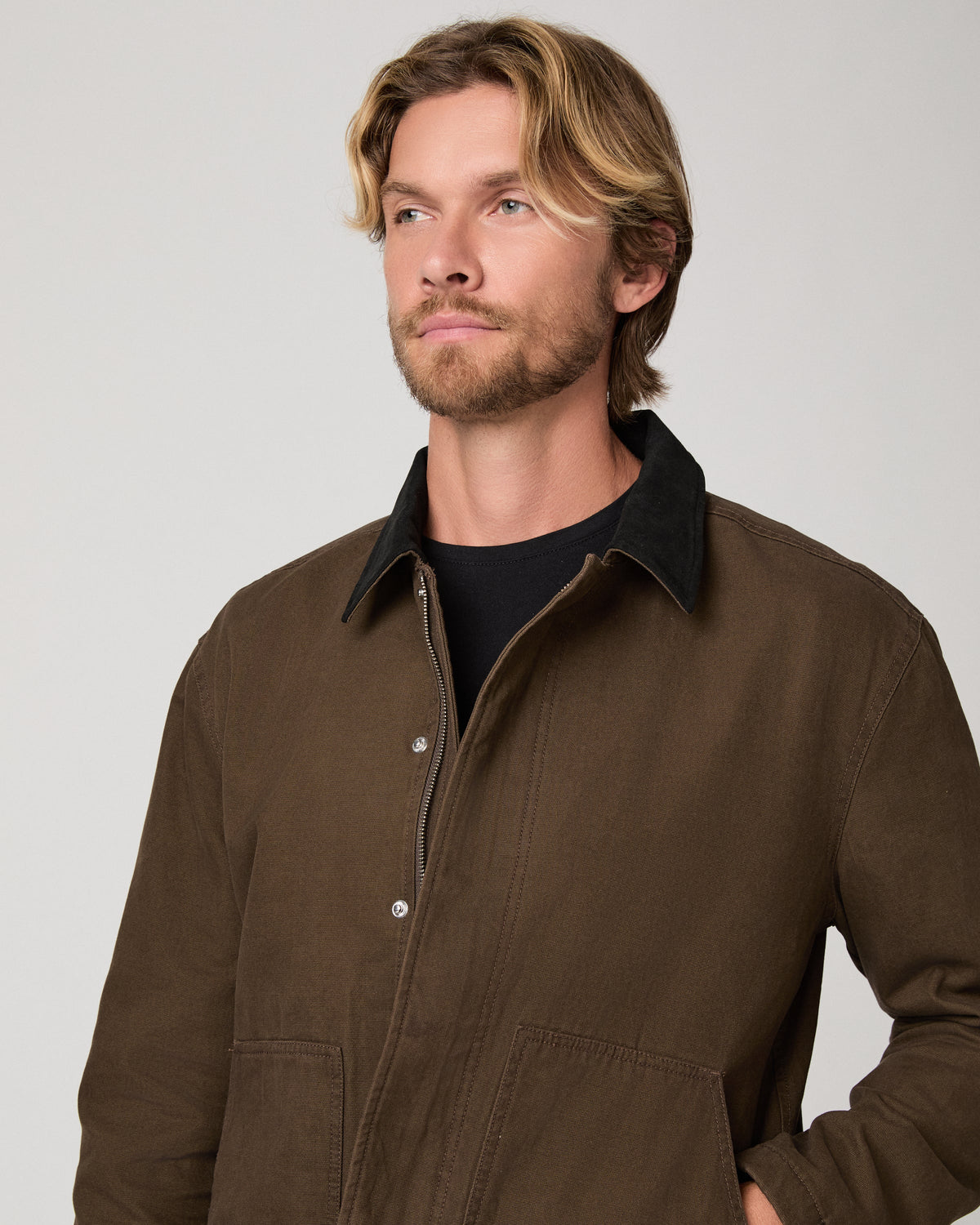 A man wearing a brown corduroy jacket with a black corduroy collar over a black t-shirt, photographed against a neutral light gray background.