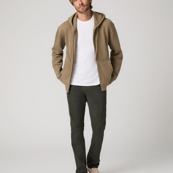 A man modeling a tan zip-up hoodie with a white t-shirt, dark gray pants, and beige slip-on shoes, standing against a light gray studio background.