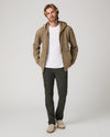 A man modeling a tan zip-up hoodie with a white t-shirt, dark gray pants, and beige slip-on shoes, standing against a light gray studio background.