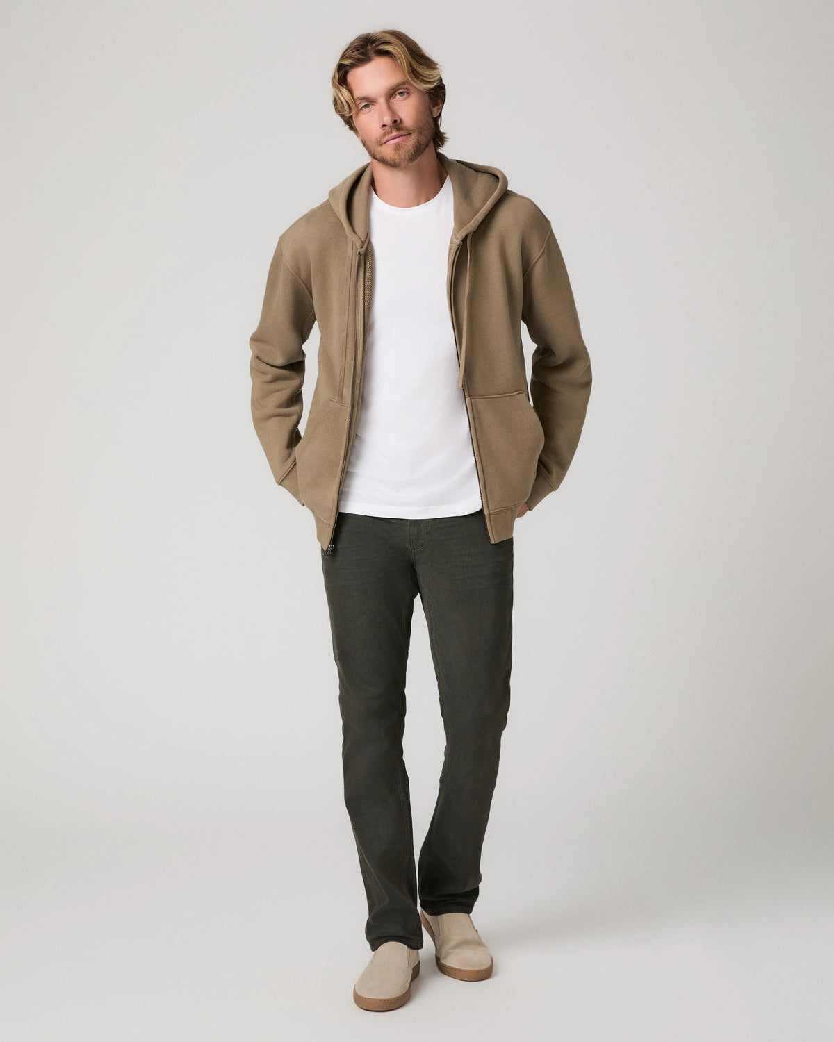 A man modeling a tan zip-up hoodie with a white t-shirt, dark gray pants, and beige slip-on shoes, standing against a light gray studio background.
