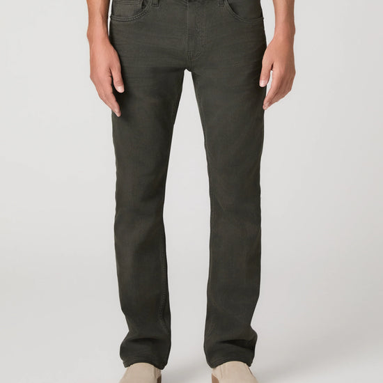 A person wearing dark olive green slim-fit jeans with a white t-shirt and beige slip-on shoes, shown from the waist down on a light gray studio background.