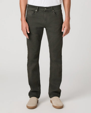 A person wearing dark olive green slim-fit jeans with a white t-shirt and beige slip-on shoes, shown from the waist down on a light gray studio background.