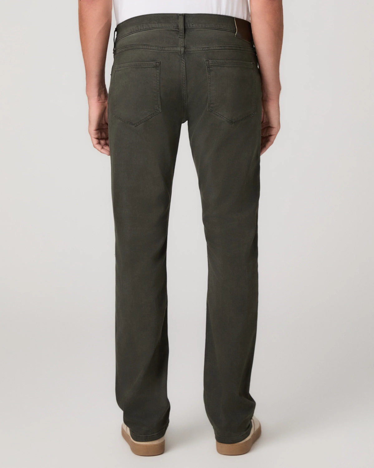 Rear view of a person wearing dark olive green straight-leg jeans with a white shirt and tan shoes, photographed against a light gray studio background.