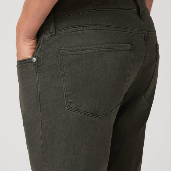 Close-up back view of a person wearing dark olive green jeans, showing the back pocket, waistband, and side pocket with a hand in the pocket, against a light gray background.