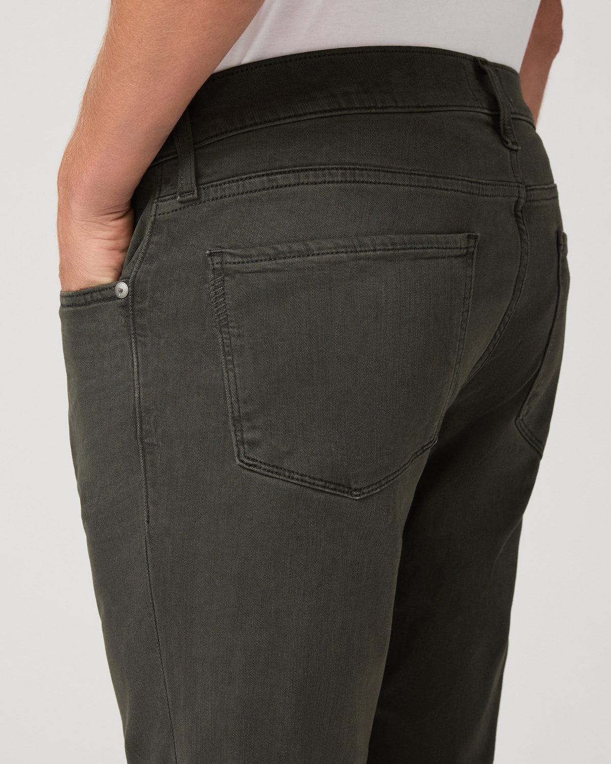 Close-up back view of a person wearing dark olive green jeans, showing the back pocket, waistband, and side pocket with a hand in the pocket, against a light gray background.