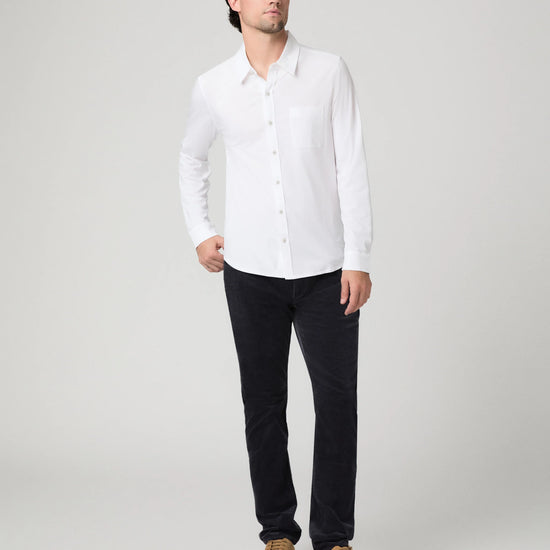 A man modeling a white long-sleeve button-down shirt with a chest pocket, paired with dark trousers and tan suede shoes, against a light gray studio background.