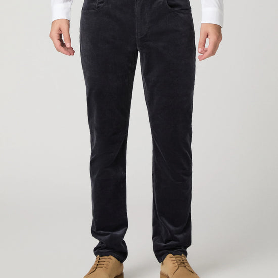 A person wearing dark navy corduroy trousers, styled with a white long-sleeve shirt and tan suede shoes, shown from the waist down on a light gray studio background.