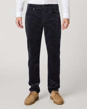 A person wearing dark navy corduroy trousers, styled with a white long-sleeve shirt and tan suede shoes, shown from the waist down on a light gray studio background.