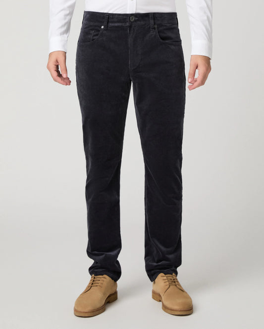 A person wearing dark navy corduroy trousers, styled with a white long-sleeve shirt and tan suede shoes, shown from the waist down on a light gray studio background.