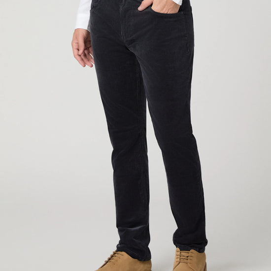 A person wearing slim-fit black corduroy trousers, styled with a white long-sleeve shirt and tan suede lace-up shoes, photographed against a light gray studio background.