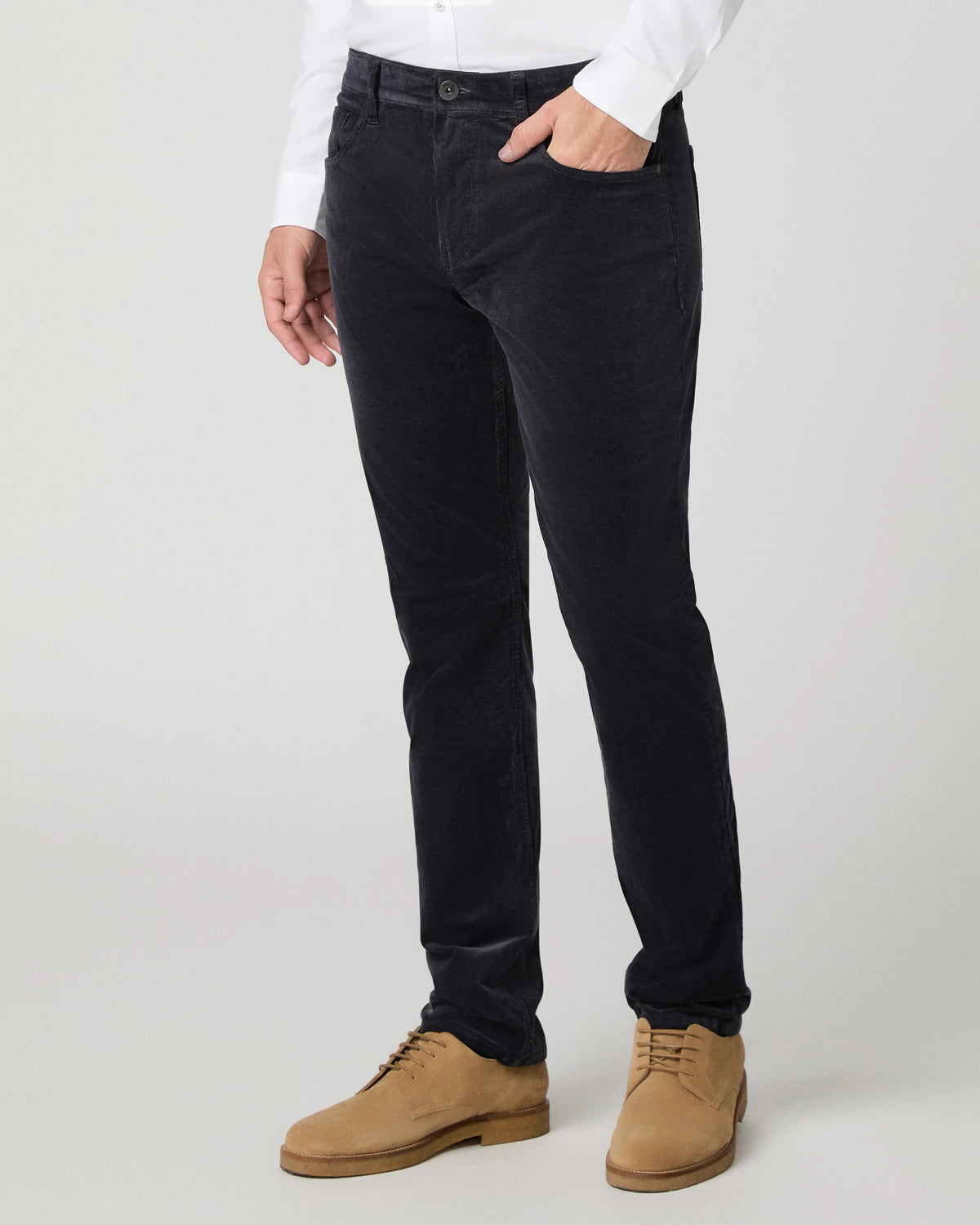A person wearing slim-fit black corduroy trousers, styled with a white long-sleeve shirt and tan suede lace-up shoes, photographed against a light gray studio background.