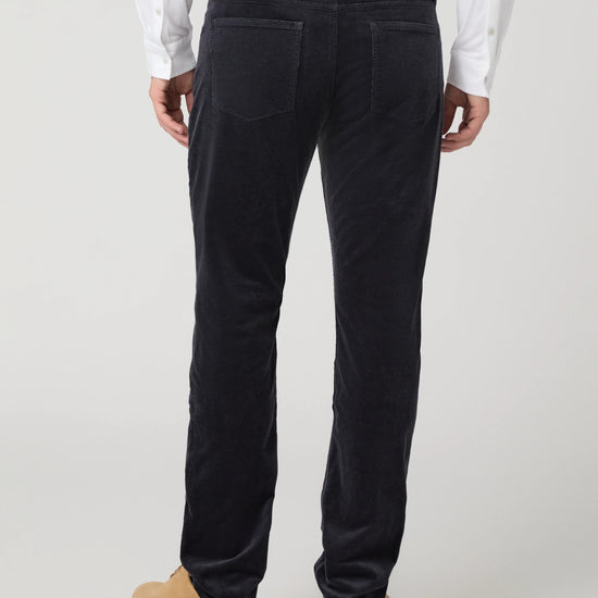 Rear view of a person wearing dark navy corduroy trousers, a white shirt, and tan boots against a light gray studio background.