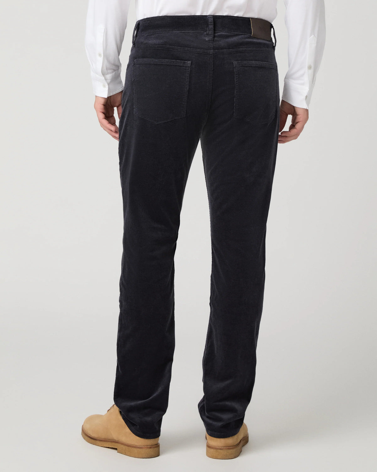 Rear view of a person wearing dark navy corduroy trousers, a white shirt, and tan boots against a light gray studio background.