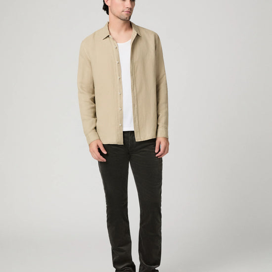 A man modeling a light beige linen button-up shirt over a white t-shirt, paired with dark charcoal pants and beige slip-on shoes, against a neutral light gray background.