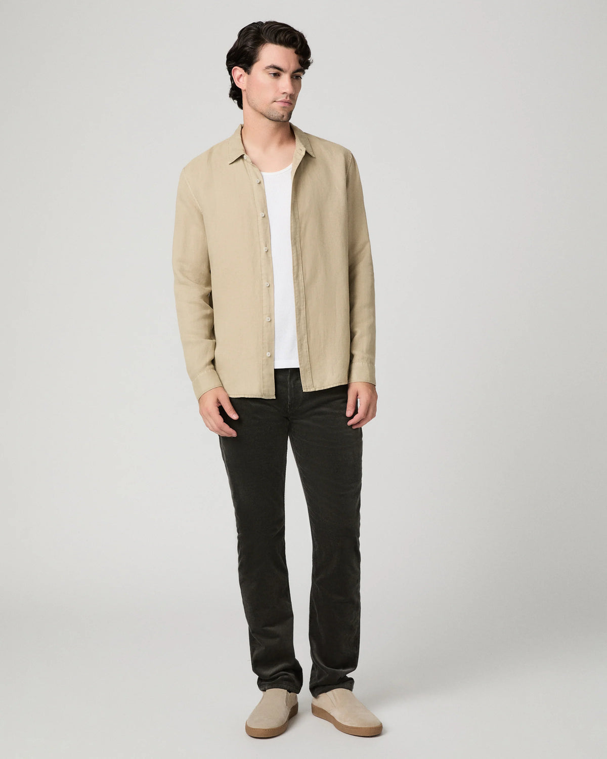 A man modeling a light beige linen button-up shirt over a white t-shirt, paired with dark charcoal pants and beige slip-on shoes, against a neutral light gray background.