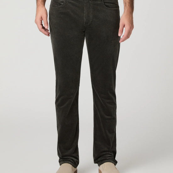 A person wearing dark charcoal corduroy trousers, styled with a white t-shirt and beige slip-on shoes, photographed against a neutral light gray studio background.