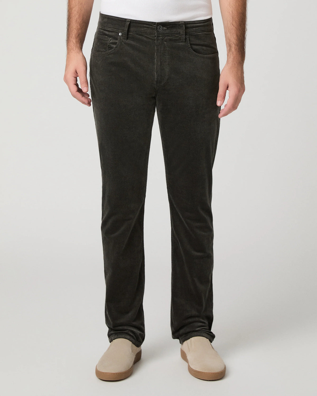 A person wearing dark charcoal corduroy trousers, styled with a white t-shirt and beige slip-on shoes, photographed against a neutral light gray studio background.