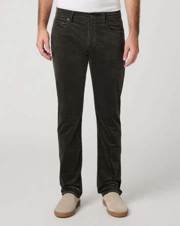 A person wearing dark charcoal corduroy trousers, styled with a white t-shirt and beige slip-on shoes, photographed against a neutral light gray studio background.