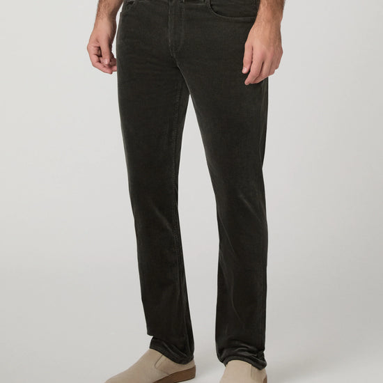 A person wearing slim-fit dark charcoal corduroy jeans with beige slip-on shoes, shown from the waist down against a light gray studio background.