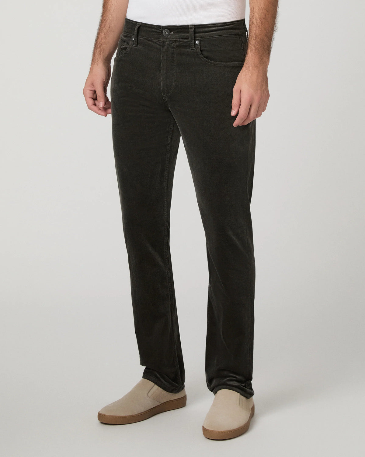 A person wearing slim-fit dark charcoal corduroy jeans with beige slip-on shoes, shown from the waist down against a light gray studio background.