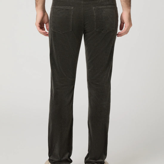 Rear view of a person wearing dark charcoal corduroy trousers, a white t-shirt, and beige platform shoes against a light gray studio background.