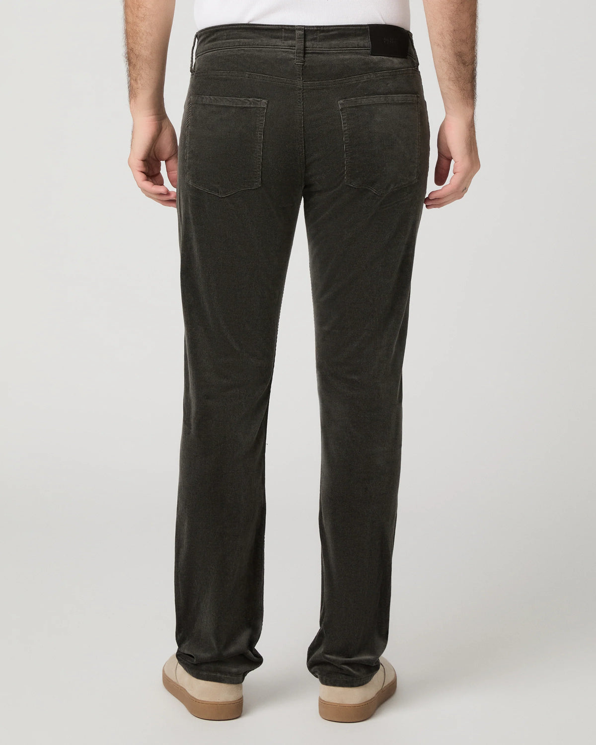 Rear view of a person wearing dark charcoal corduroy trousers, a white t-shirt, and beige platform shoes against a light gray studio background.