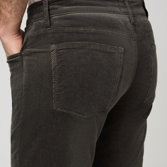 Close-up back view of a person wearing dark charcoal corduroy pants, showing the back pockets and waistband against a light gray studio background.