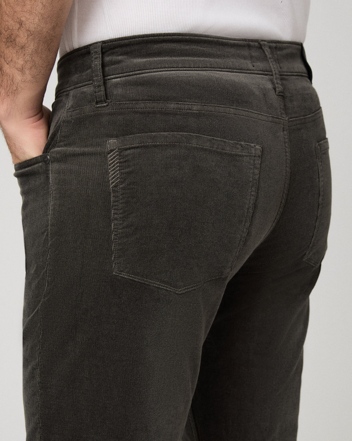 Close-up back view of a person wearing dark charcoal corduroy pants, showing the back pockets and waistband against a light gray studio background.