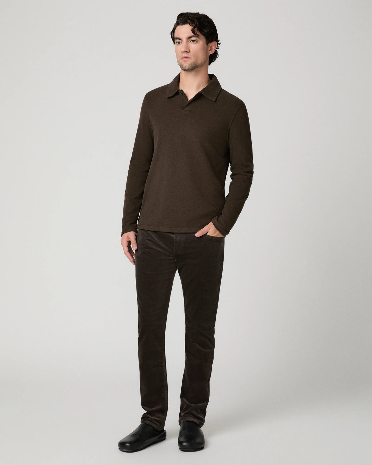 A man modeling a dark brown long-sleeve polo shirt and matching dark brown corduroy trousers against a light gray studio background.