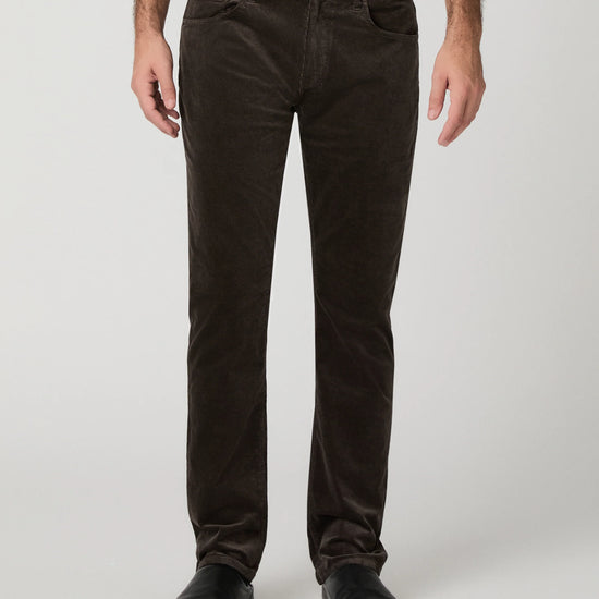A person wearing dark brown corduroy trousers, a white shirt, and black shoes, standing against a light gray studio background.