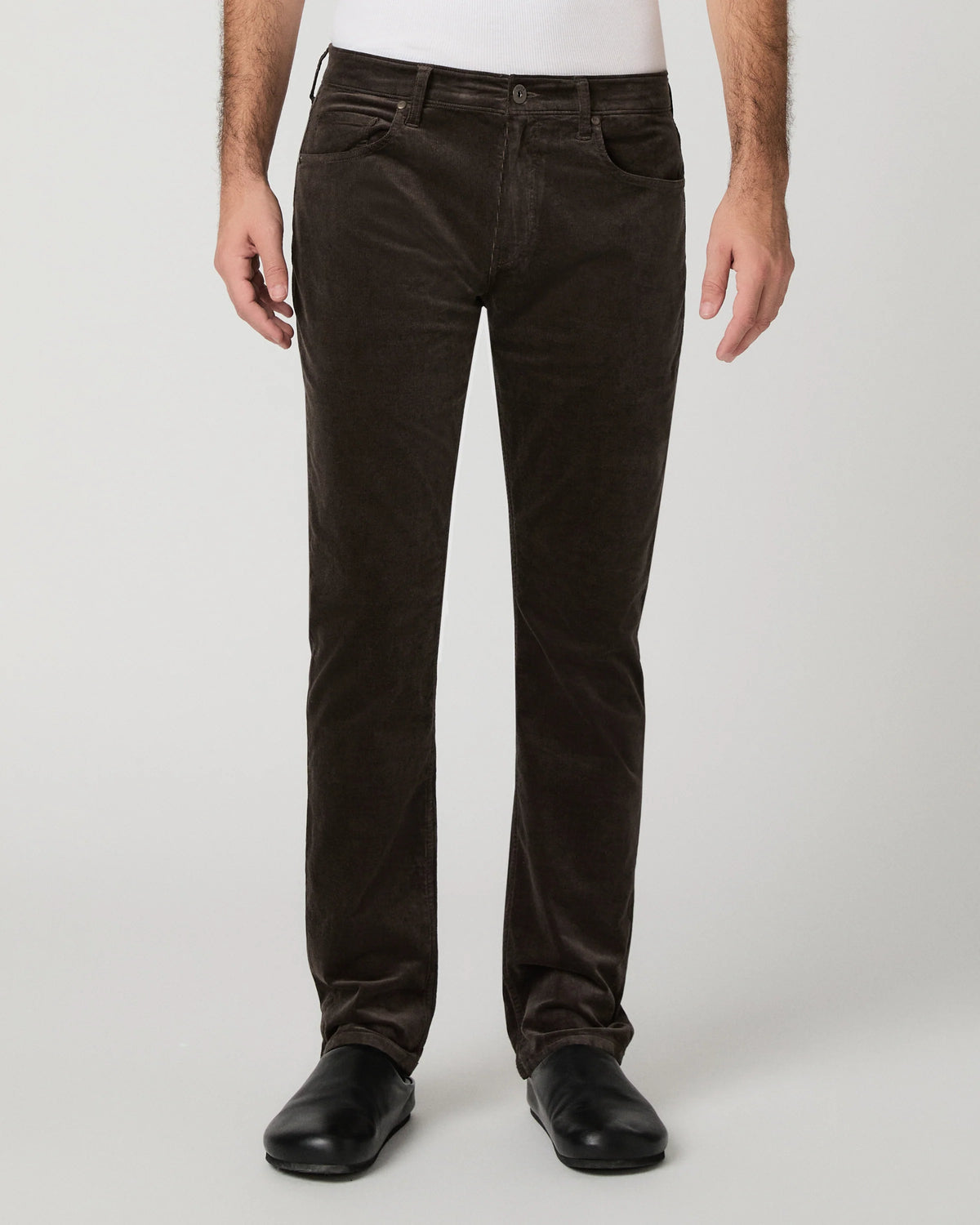 A person wearing dark brown corduroy trousers, a white shirt, and black shoes, standing against a light gray studio background.