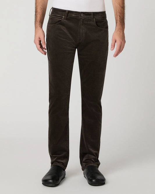 A person wearing dark brown corduroy trousers, a white shirt, and black shoes, standing against a light gray studio background.