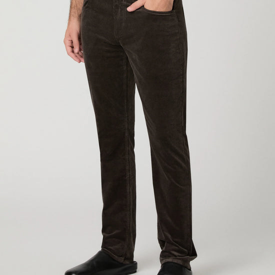 A person wearing dark brown corduroy trousers, styled with a white shirt and black shoes, shown from the waist down against a light gray studio background.