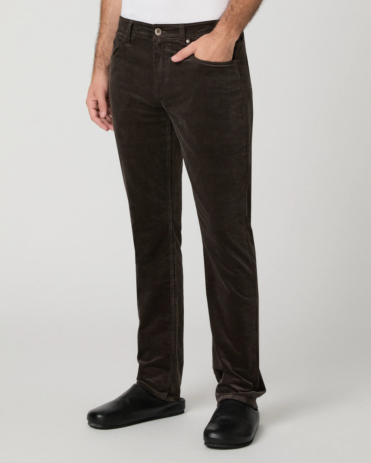 A person wearing dark brown corduroy trousers, styled with a white shirt and black shoes, shown from the waist down against a light gray studio background.