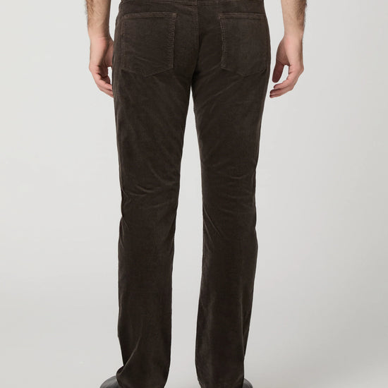 Rear view of a person wearing dark brown corduroy trousers, a white shirt, and black shoes against a light gray studio background.