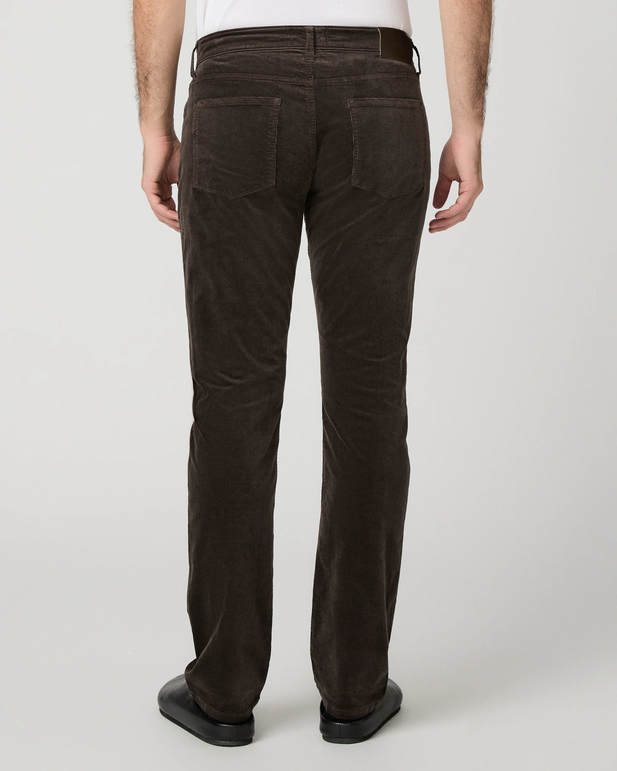 Rear view of a person wearing dark brown corduroy trousers, a white shirt, and black shoes against a light gray studio background.