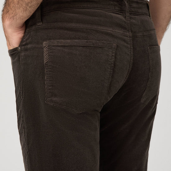 Close-up back view of a person wearing dark brown corduroy trousers, showing the back pocket and waistband against a light gray background.