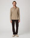 A man modeling a beige long-sleeve knit polo shirt with dark brown trousers and beige slip-on shoes, standing against a light gray studio background.