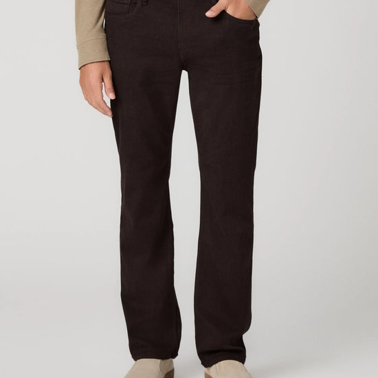 A person wearing dark brown straight-leg jeans, a beige long-sleeve top, and beige slip-on shoes, standing against a light gray studio background.