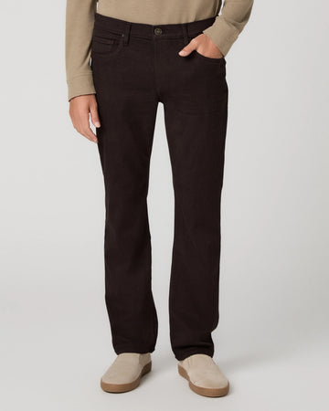 A person wearing dark brown straight-leg jeans, a beige long-sleeve top, and beige slip-on shoes, standing against a light gray studio background.