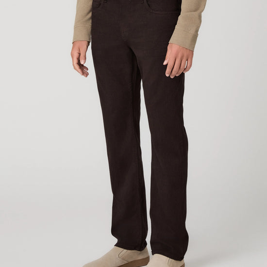 A person wearing dark brown slim-fit jeans, a beige long-sleeve top, and beige slip-on shoes, standing against a light gray studio background.