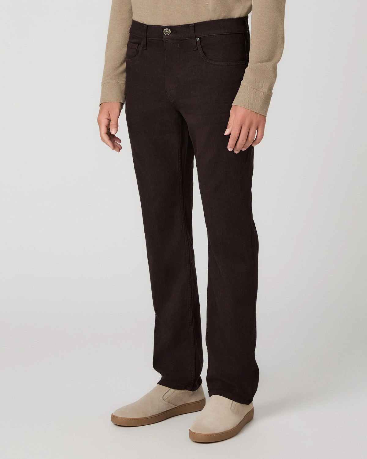 A person wearing dark brown slim-fit jeans, a beige long-sleeve top, and beige slip-on shoes, standing against a light gray studio background.