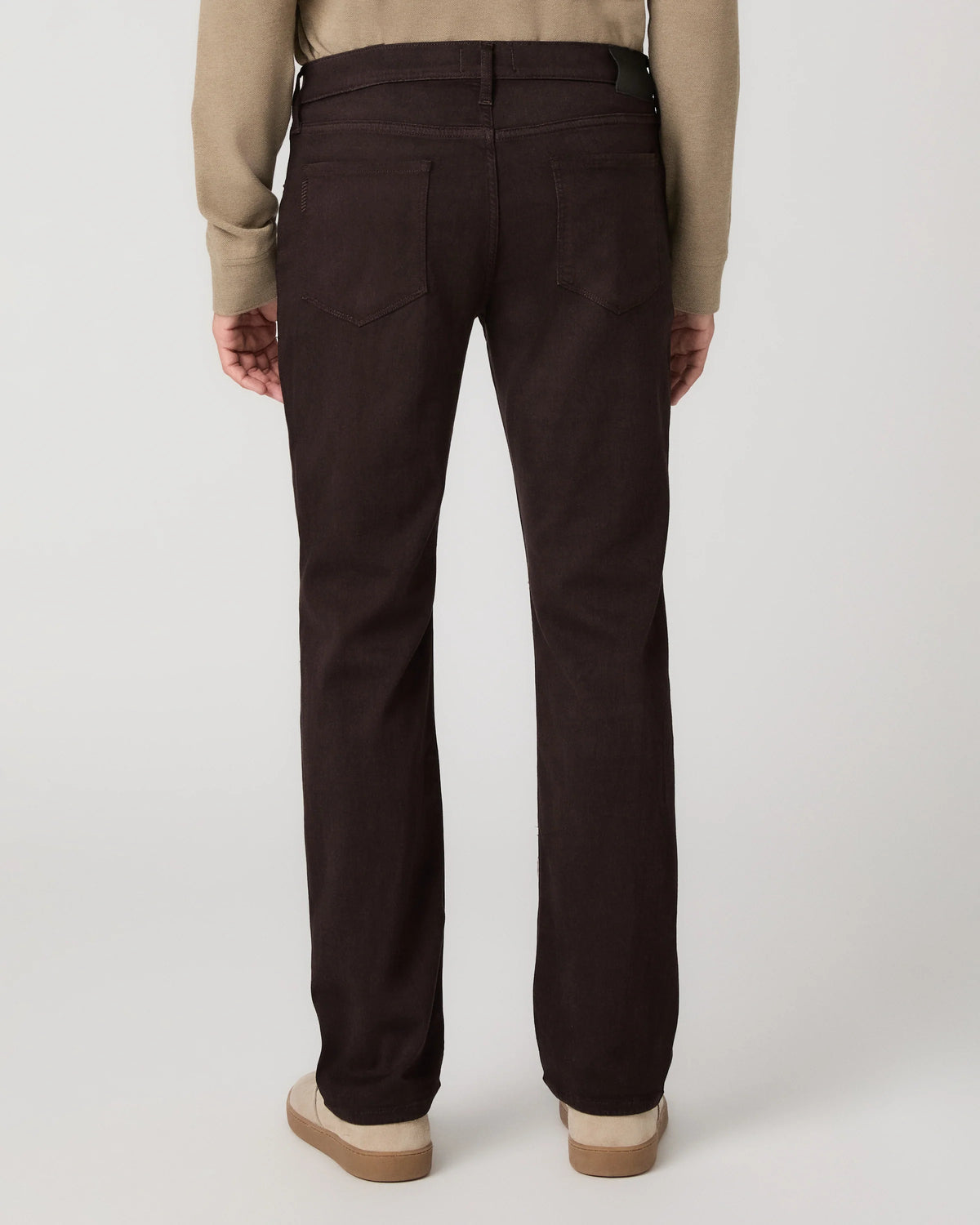 Rear view of a person wearing dark brown straight-leg jeans with a beige long-sleeve top and beige platform shoes, against a light gray studio background.