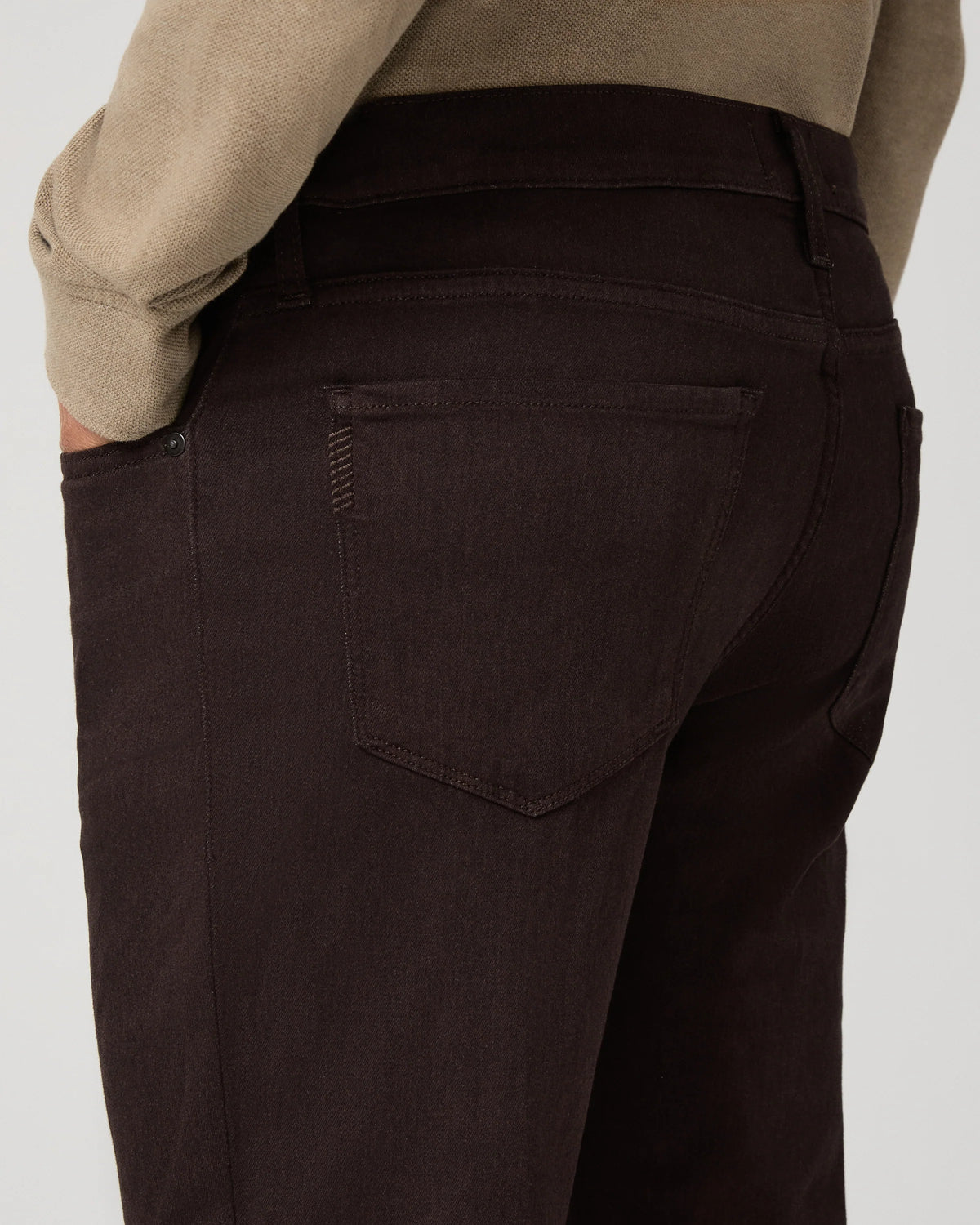 Close-up back view of a person wearing dark brown or very deep espresso corduroy trousers, paired with a beige sweater, against a light gray studio background.